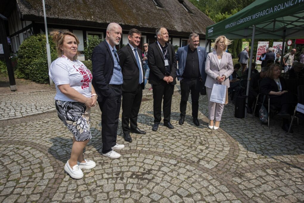 Grupa uczestników wydarzenia stoi na dziedzińcu przed budynkiem, słuchając wystąpienia organizatorów (kliknięcie powiększy zdjęcie).