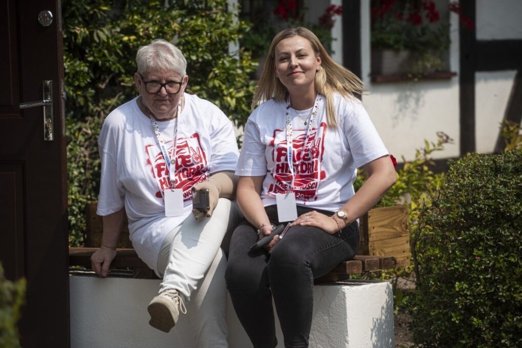 Dwie kobiety w koszulkach z logo festiwalu siedzą na ławce przed budynkiem (kliknięcie powiększy zdjęcie).