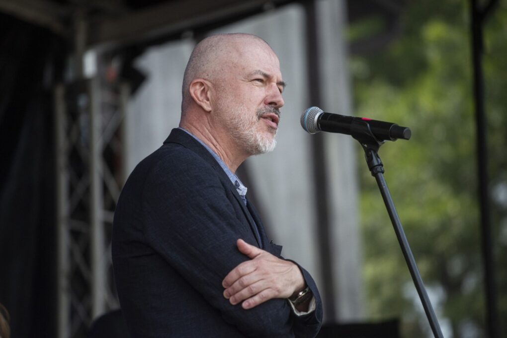 Prelegent przemawia do mikrofonu na scenie plenerowej podczas wydarzenia (kliknięcie powiększy zdjęcie).