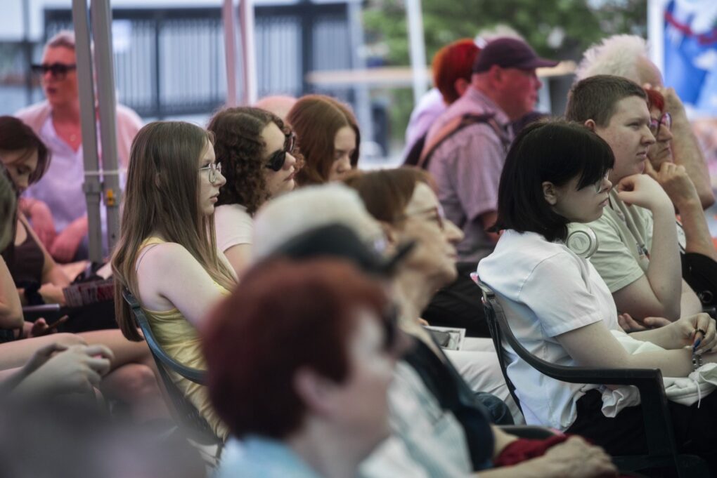 Uczestnicy wydarzenia siedzą w rzędach i słuchają programu na scenie (kliknięcie powiększy zdjęcie).