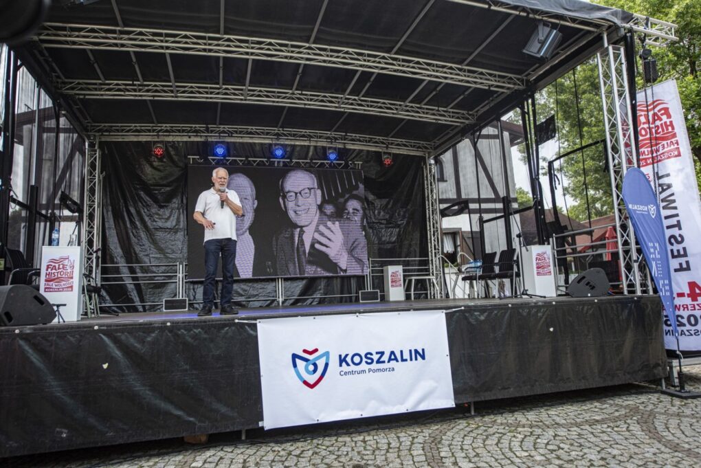 Scena plenerowa z prowadzącym wystąpienie oraz ekranem z archiwalnym zdjęciem w tle (kliknięcie powiększy zdjęcie).