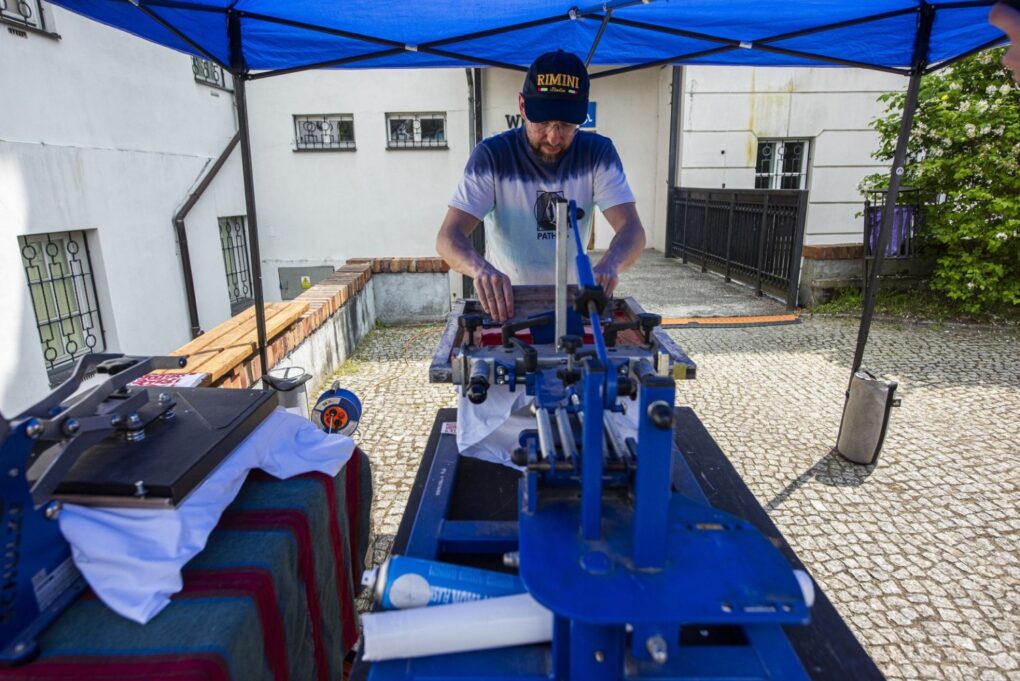 Mężczyzna obsługuje urządzenie do nadruku koszulek podczas warsztatów na stoisku plenerowym (kliknięcie powiększy zdjęcie).