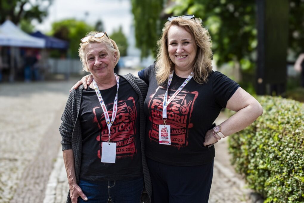 Dwie kobiety w czarnych koszulkach z czerwonym logo festiwalu stoją razem na dziedzińcu (kliknięcie powiększy zdjęcie).