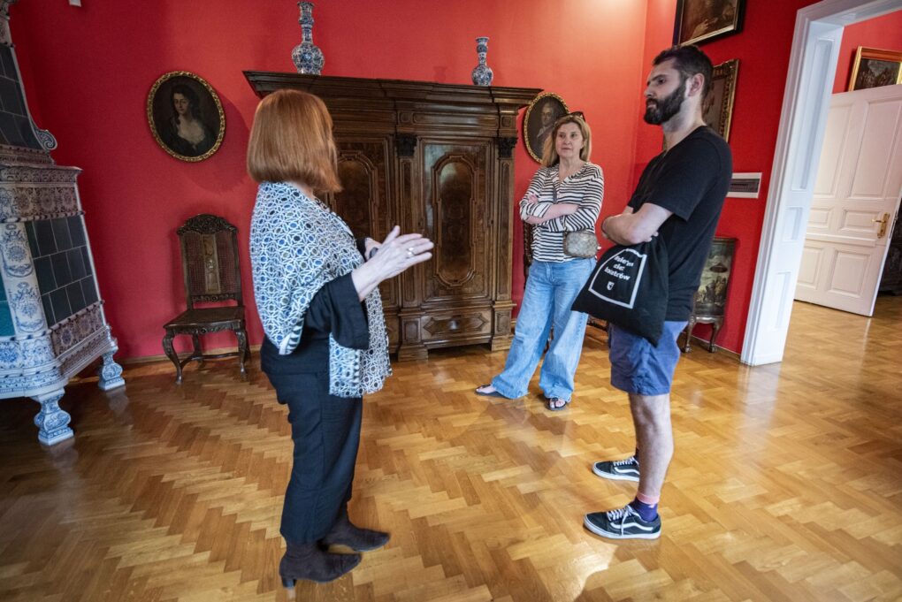 Zwiedzający rozmawiają w sali muzealnej przy zabytkowych meblach i eksponatach (kliknięcie powiększy zdjęcie).