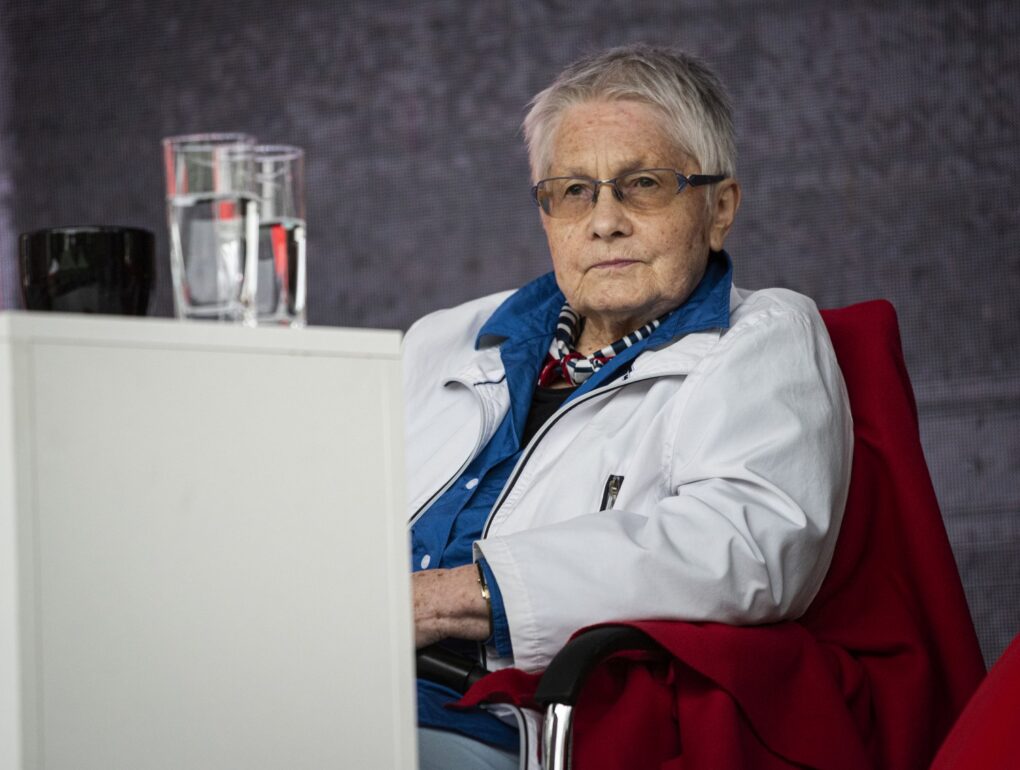 Starsza kobieta siedzi na scenie przy stole podczas panelu dyskusyjnego (kliknięcie powiększy zdjęcie).