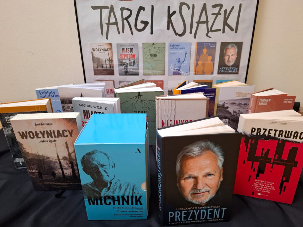 Na stole stoją rozłożone książki. Na pierwszym planie publikacja Aleksandra Kwaśniewskiego i Adama Michnika. W tle, na ścianie, wisi plakat promujący Targi Książki Historycznej i Regionalnej (kliknięcie spowoduje powiększenie zdjęcia).