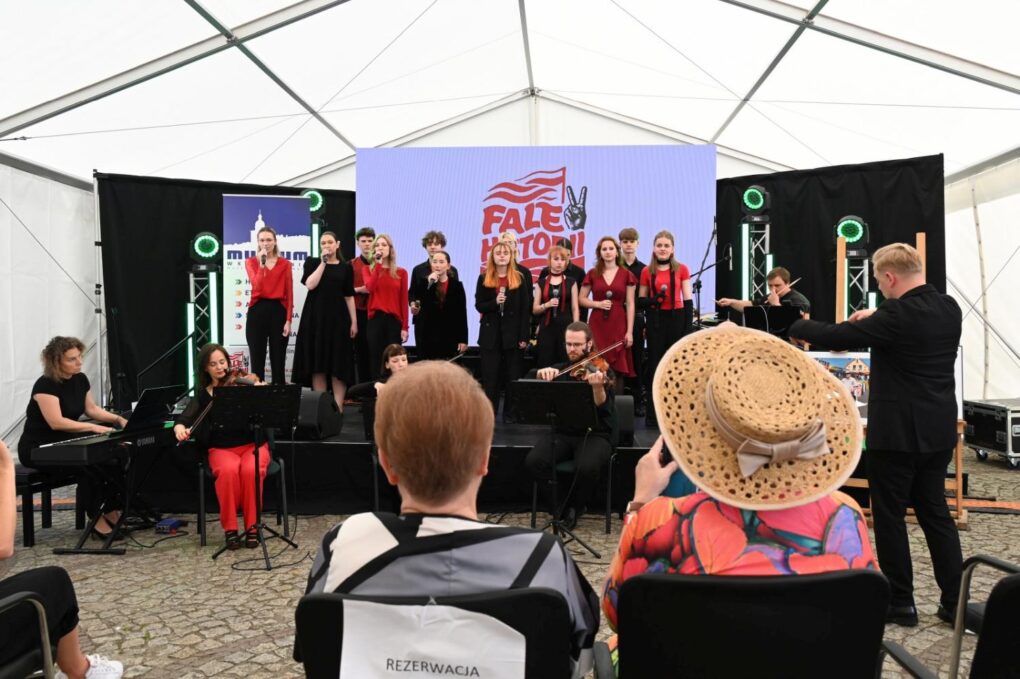 Młodzieżowy chór i zespół instrumentalny występują na scenie podczas festiwalu „Fale Historii”. Dyrygent prowadzi wykonawców, a publiczność przysłuchuje się koncertowi (kliknięcie powiększy zdjęcie).