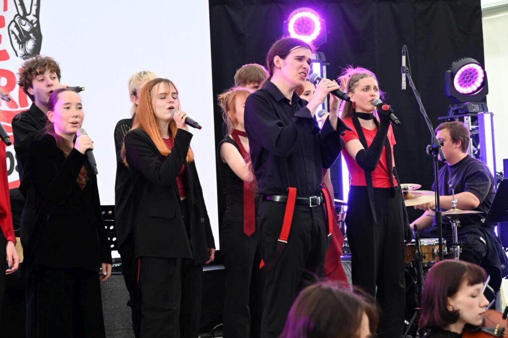 Młodzi wokaliści wykonują utwór podczas koncertu w ramach festiwalu „Fale Historii”. Na pierwszym planie soliści śpiewają do mikrofonów, a w tle akompaniuje zespół muzyczny (kliknięcie powiększy zdjęcie).